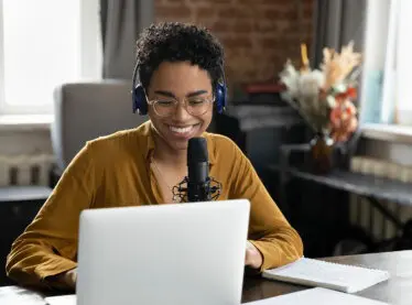 Smiling woman wearing wireless headphones, talking into a microphone, and looking at a laptop screen.