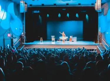 An actor on a blue stage giving a performance in front of a packed house.