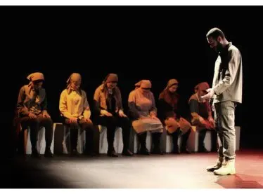 Actors on stage performing a play. One recites a monologues, others sit in silence.