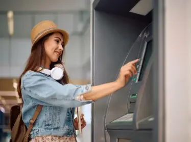 Asian woman in a sun hat and denim jacket withdrawing money from an ATM.