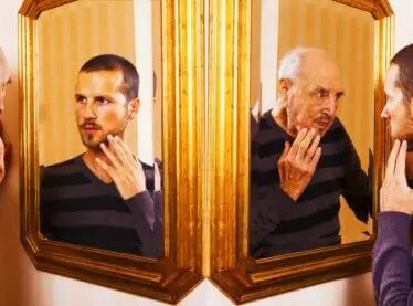 Two actors looking in the mirrors at their old and young reflections.