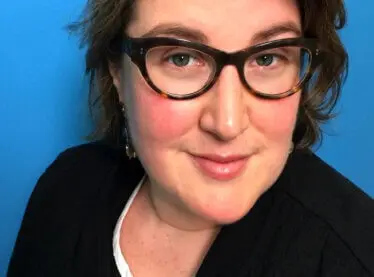 Casting Director Julie Schubert smiling in front of a blue background, wearing a black shirt.