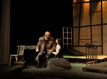 Two actors in vintage clothing performing in a play surrounded by chairs and wood pallets.