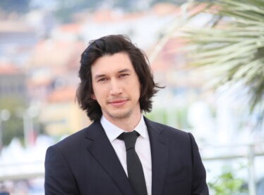 Adam Driver smiling outside in a suit.