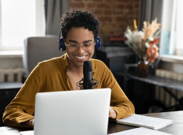 Smiling woman wearing wireless headphones, talking into a microphone, and looking at a laptop screen.