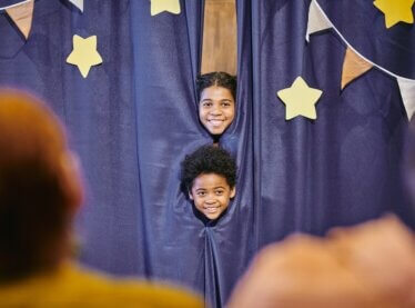 Two children smiling behind a purple curtain as parents watch their play.