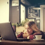 A blonde woman at a coffee shop watching an entertaining video on her laptop.