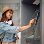 Asian woman in a sun hat and denim jacket withdrawing money from an ATM.