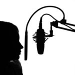 Silhouette of a femaile voice actor and a microphone on a stand in front of her as she records.