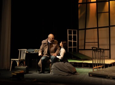 Two actors in vintage clothing performing in a play surrounded by chairs and wood pallets.