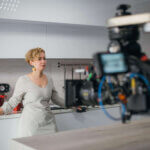 Actress in a kitchen filming a commercial, camera recording in the middle right.